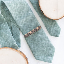 Load image into Gallery viewer, A pine tree brown and black pattern brass tie clip is styled with a sage green tie against a white background with wooden display props.