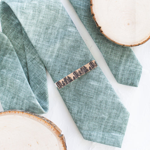 A pine tree brown and black pattern brass tie clip is styled with a sage green tie against a white background with wooden display props.