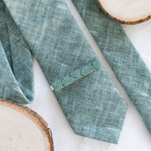 Load image into Gallery viewer, A leaf pattern brass tie clip is styled with a sage green tie against a white background with wooden display props.