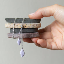 Load image into Gallery viewer, A hand holds a stack of flat rocks to display a silver chain bolo necklace with two lilac petals hanging from the ends.
