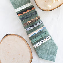 Load image into Gallery viewer, A collection of nature-inspired polymer clay brass tie clips are arranged on a sage green tie, with a white textured background and wooden display props.