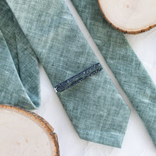Load image into Gallery viewer, A starry night sky pattern brass tie clip is styled with a sage green tie against a white background with wooden display props.