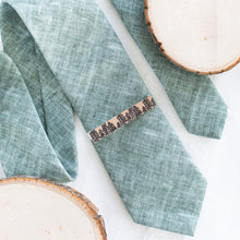 Load image into Gallery viewer, A pine tree brown and black pattern brass tie clip is styled with a sage green tie against a white background with wooden display props.

