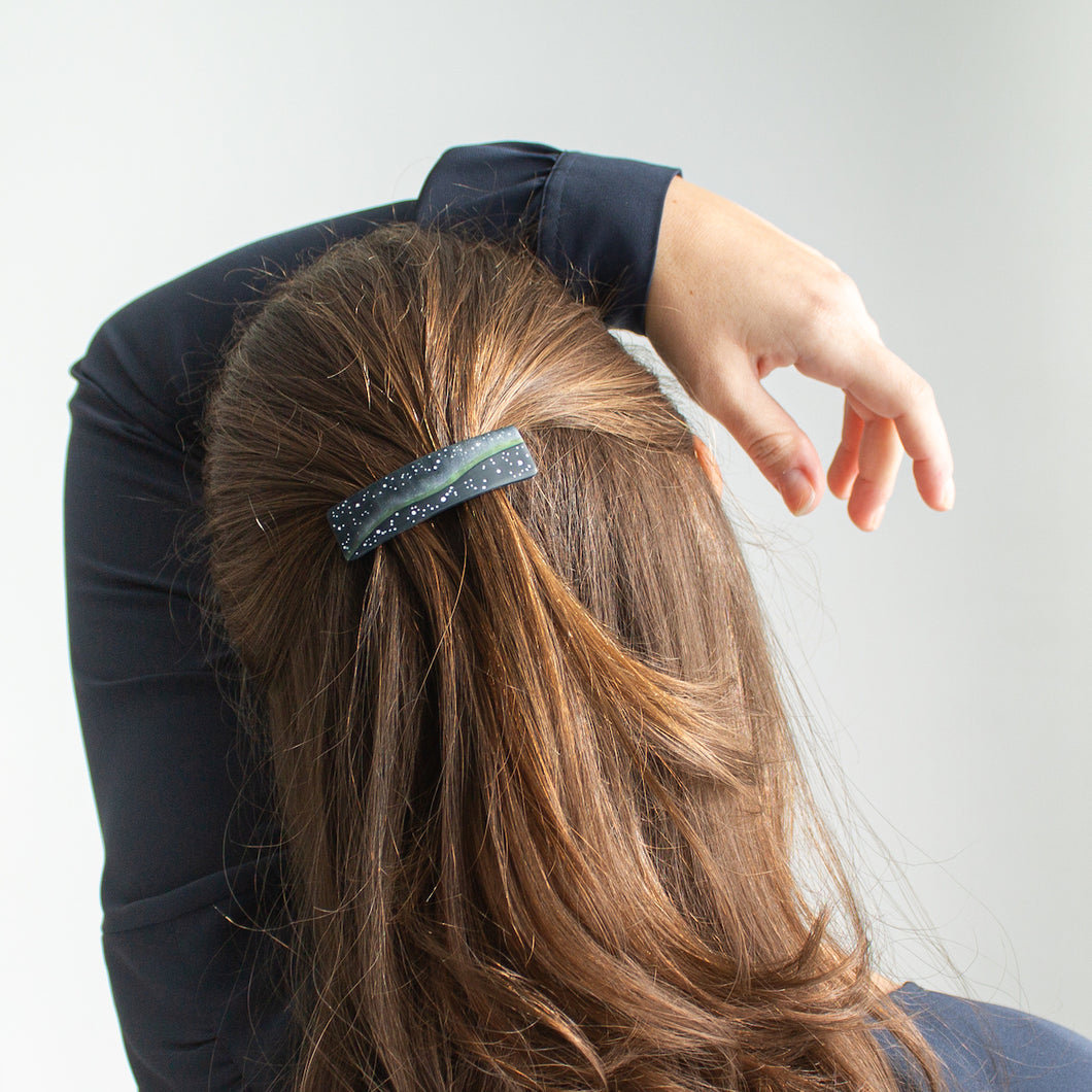 A model with brown hair wears a small dark blue polymer clay barrette that has a northern lights design.
