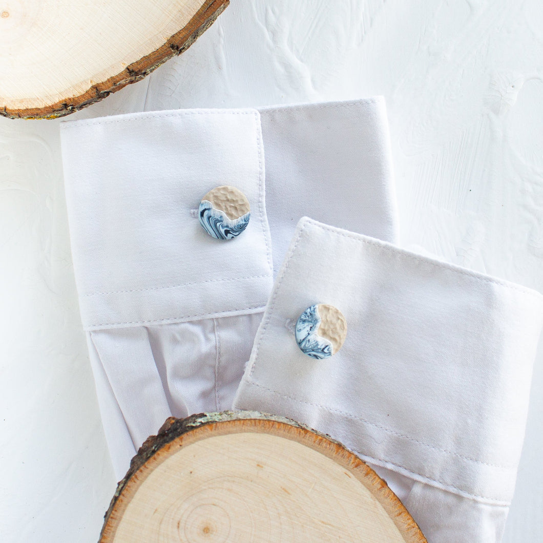 Cufflinks with a blue and beige wave and beach pattern are styled on the cuffs of a white shirt, against a white background.