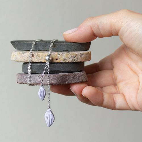 A hand holds a stack of flat rocks to display a silver chain bolo necklace with two lilac petals hanging from the ends.