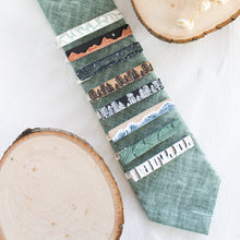 Load image into Gallery viewer, A collection of nature-inspired polymer clay brass tie clips are arranged on a sage green tie, with a white textured background and wooden display props.

