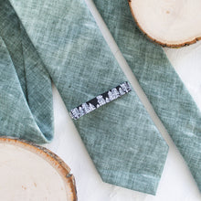Load image into Gallery viewer, A pine tree black and white pattern brass tie clip is styled with a sage green tie against a white background with wooden display props.
