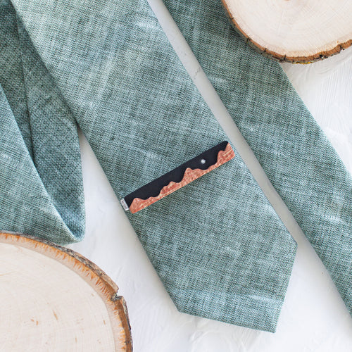 A red rock landscape pattern brass tie clip is styled with a sage green tie against a white background with wooden display props.