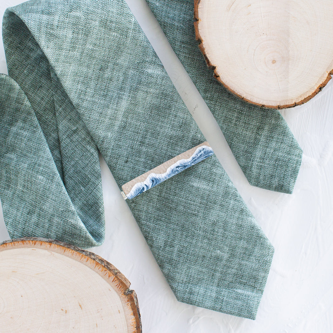 A waves and sand pattern brass tie clip is styled with a sage green tie against a white background with wooden display props.