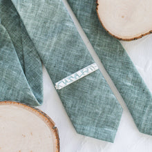 Load image into Gallery viewer, A glacier-inspired pattern brass tie clip is styled with a sage green tie against a white background with wooden display props.
