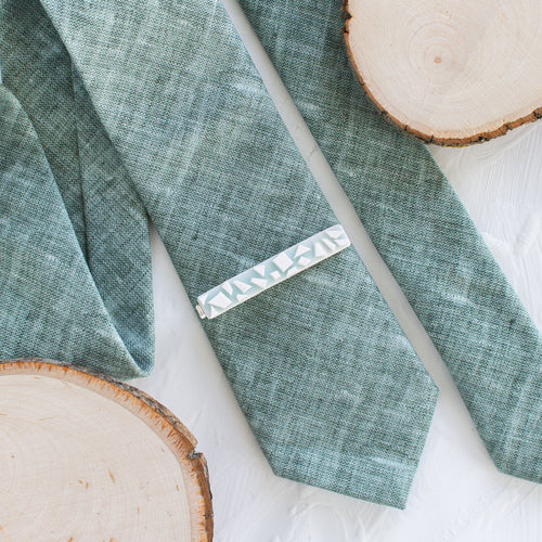 A glacier-inspired pattern brass tie clip is styled with a sage green tie against a white background with wooden display props.